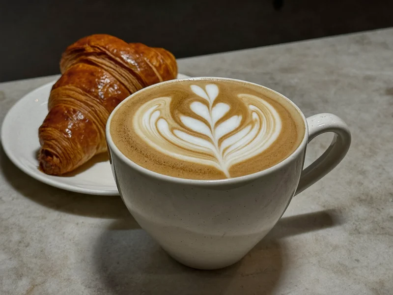 Flat white cu latte art, alături de un croissant mic, pe masă de piatră.
